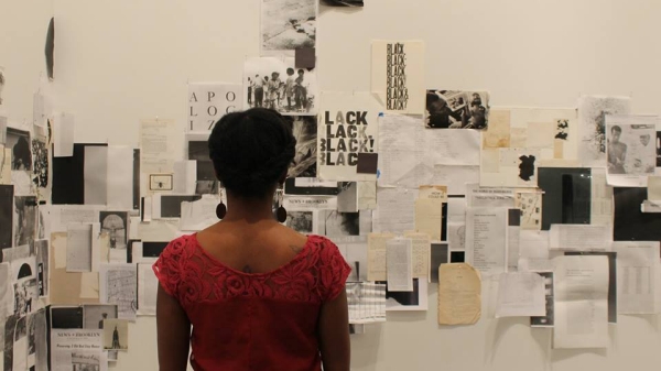 Black woman wearing red shirt, back to audience, looks at papers taped to a white wall